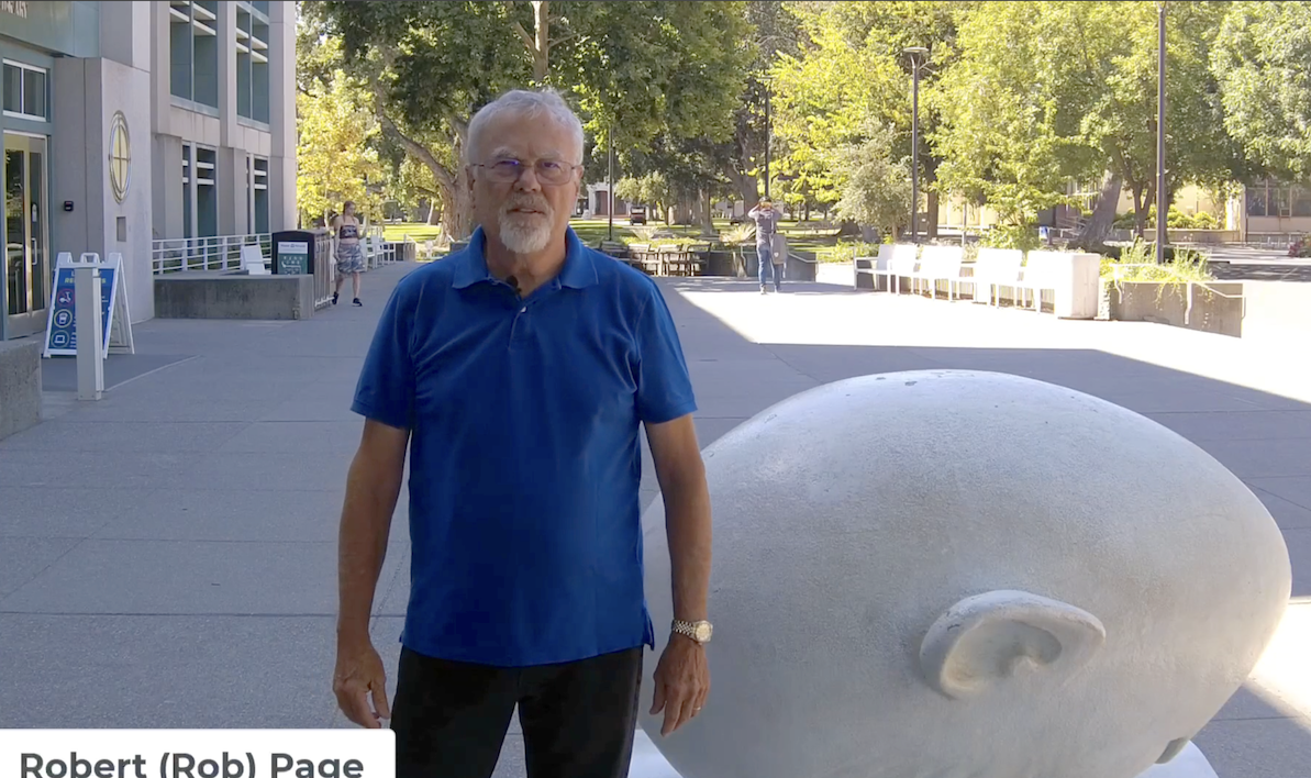 Rob Page in blue polo standing by large white spiral sculpture on sunny plaza