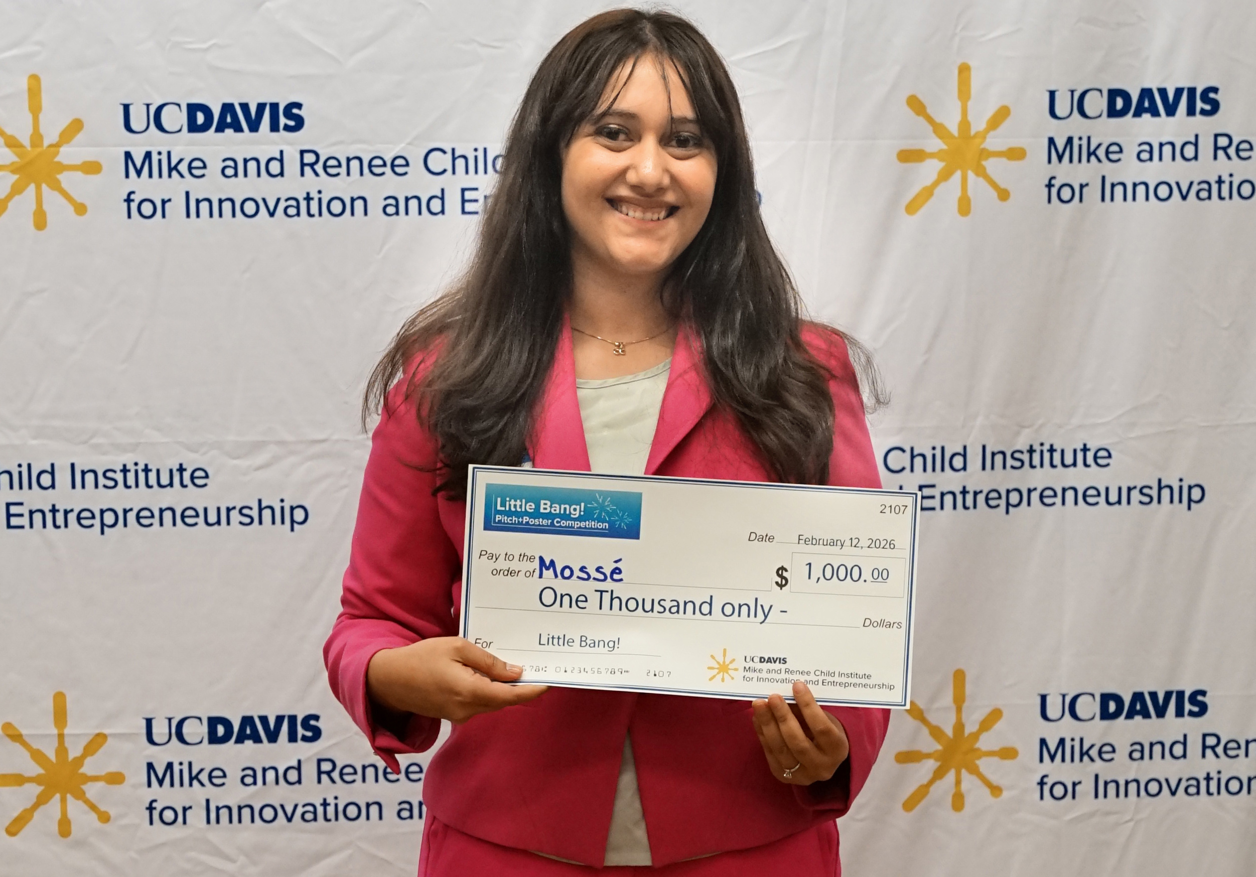 Smiling woman in a pink blazer holding a large check in front of a branded backdrop.