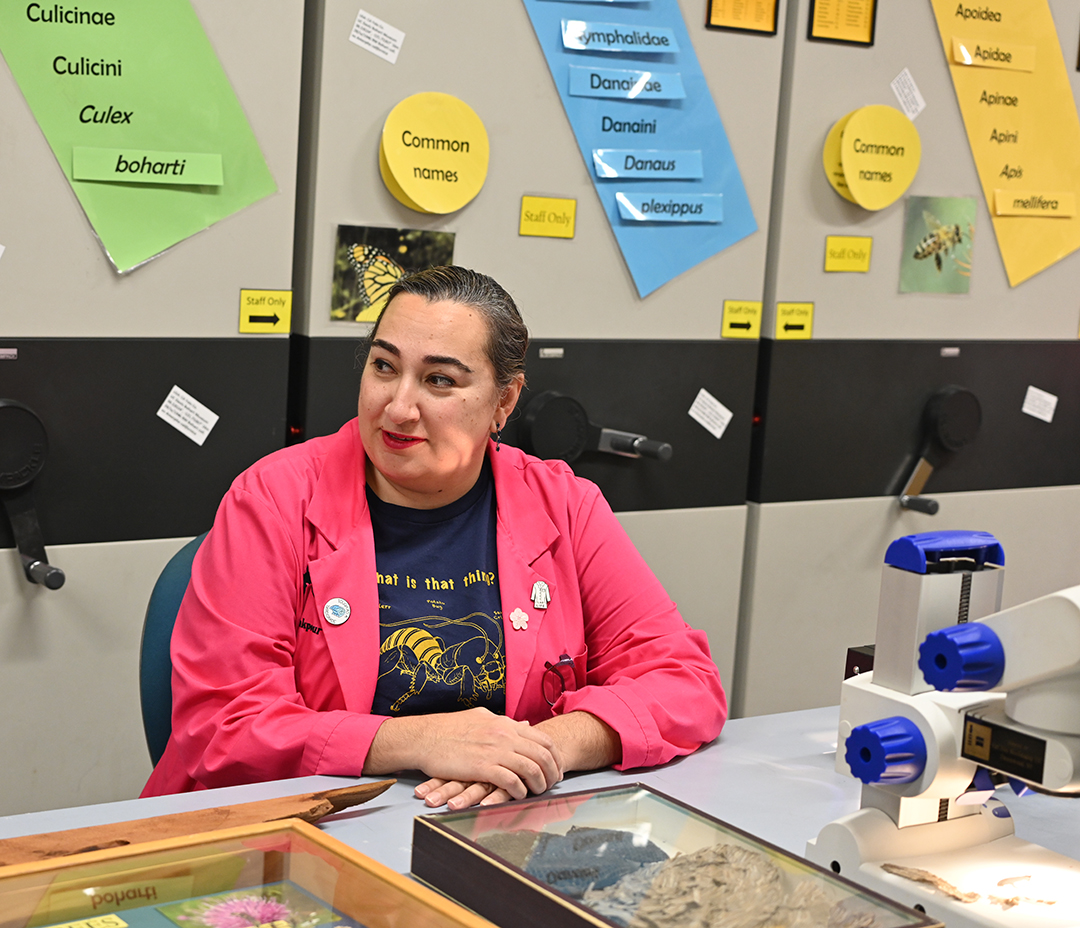 A person in a pink lab coat sits at a table with scientific equipment and displays behind.
