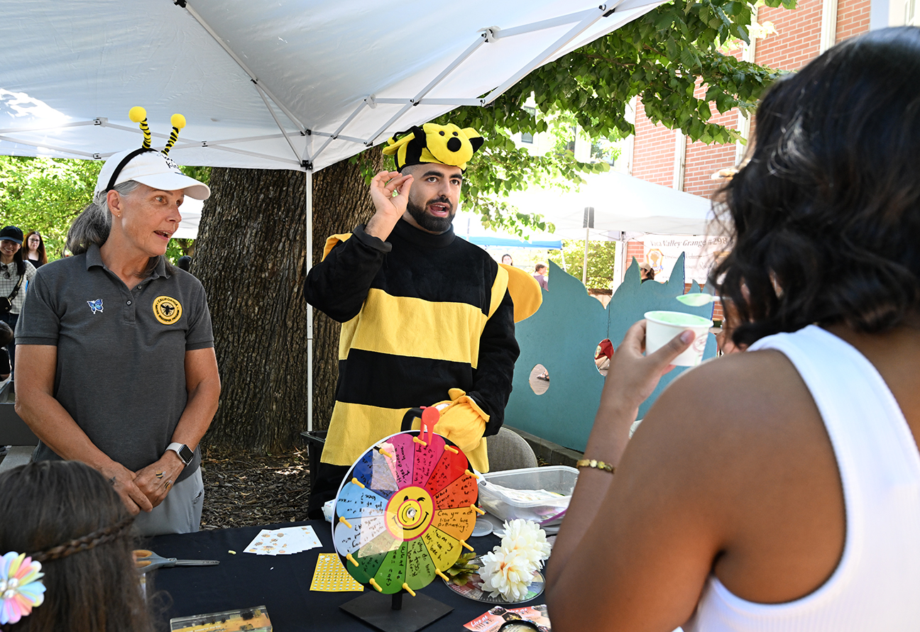 Man in bumblebee costume tasting honey at outdoor vendor booth, colorful crafts on table