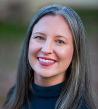 Smiling woman with long, straight gray hair, wearing a black turtleneck in a blurred green background.