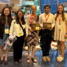 Eight smiling people wearing conference badges stand in a convention lobby with booths